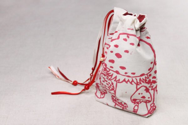 Closed standing cream handprinted fabric drawstring bag with spotted red white mushroom print with ribbons and beads to side
