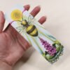 Artist holding Bookmark of Illustration “Sacro Nectare” featuring a honey bee with sun and foxglove plant (digitalis)