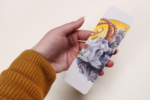 Artist holding Bookmark of Illustration “Mountain Goat Elder” featuring a mountain goat with the sun and edelweiss flowers